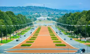 Widok na Parliament House, symbol tego, co jest stolicą Australii. Długa aleja z drzewami i samochodami prowadzi do budynku.