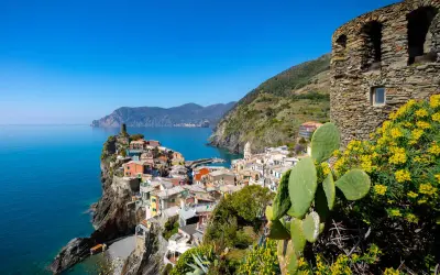 Kolorowe domy Cinque Terre wznoszą się na klifie nad turkusowym morzem. W oddali widać góry, a na pierwszym planie kaktus i żółte kwiaty.