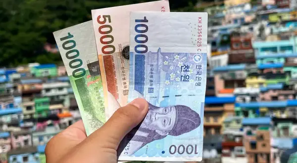 Dłoń trzyma banknoty wonów: 10000, 5000 i 1000. Tło to kolorowe domy, sugerujące pytanie: ile kosztuje wyjazd do Korei Południowej?