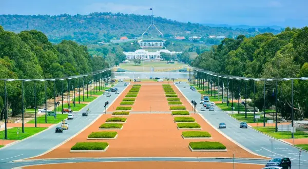 Widok na Parliament House, symbol tego, co jest stolicą Australii. Długa aleja z drzewami i samochodami prowadzi do budynku.
