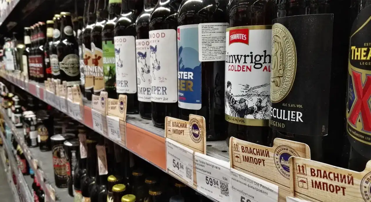 Rząd butelek piwa na półce sklepowej, niektóre z etykietami w języku ukraińskim, sugerującymi import. Widać ceny i nazwy piw, np. "Thwaites Wainwright Golden Ale".