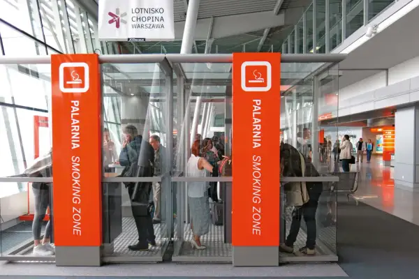 Szklane kabiny palarni na lotnisku z pomarańczowymi oznaczeniami i ludźmi w środku.