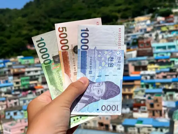 Dłoń trzyma banknoty wonów: 10000, 5000 i 1000. Tło to kolorowe domy, sugerujące pytanie: ile kosztuje wyjazd do Korei Południowej?